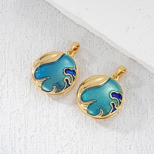 Op de afbeelding: Twee goudkleurige hangers met een rond, oceaan-thema ontwerp. Elke hanger heeft een gouden rand, een turquoise-blauwe emaille achtergrond en een donkerblauw golfpatroon. De hangers hebben een kleine lus voor een ketting.