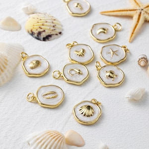 May include: Gold-coloured pendants with white enamel and marine designs, such as shells, starfish, and a turtle. The pendants are displayed on a white surface, alongside seashells and a starfish.