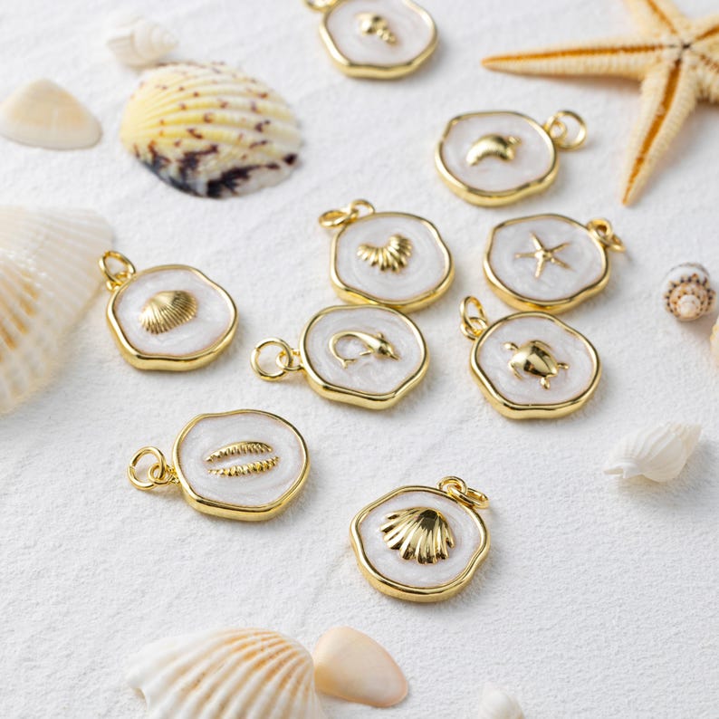 May include: Gold-coloured pendants with white enamel and marine designs, such as shells, starfish, and a turtle. The pendants are displayed on a white surface, alongside seashells and a starfish.