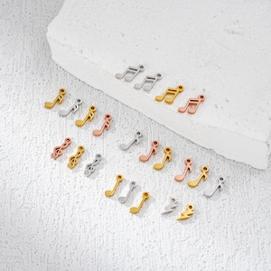 Peut inclure: Une collection de petits charms en forme de notes de musique métalliques, en or, argent et or rose. Les breloques sont disposées sur une surface texturée blanc cassé, certaines sur un bloc blanc. Divers modèles de notes de musique.