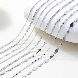 Puede incluir: Una variedad de collares plateados con varios estilos de cadena. Las cadenas incluyen diseños de eslabones delicados, cuentas y cruces, exhibidos sobre un fondo blanco. Los collares están dispuestos diagonalmente.