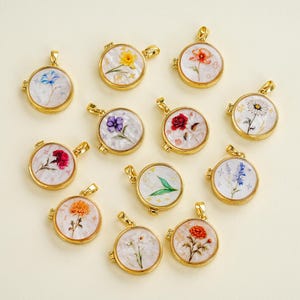 Op de afbeelding: Een verzameling goudkleurige, ronde hangers, elk met een ander ontwerp van geperste bloemen. De bloemen zijn geplaatst tegen een witte achtergrond en ingekapseld in heldere hars. De hangers hebben een scharnierend ontwerp en een kleine lus voor een ketting.