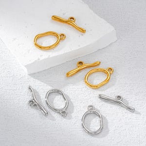 以下が含まれることがあります： 金と銀色のジュエリークラスプとリング。クラスプは、両端に小さなループが付いた湾曲したバーのデザインです。リングはループ付きの楕円形です。ジュエリーは白い表面に表示されています。