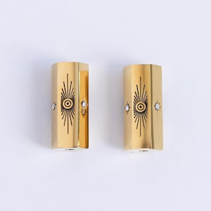 May include: A pair of gold-toned earrings with a starburst design and a small eye in the center. Each earring has a small clear rhinestone on either side of the eye.