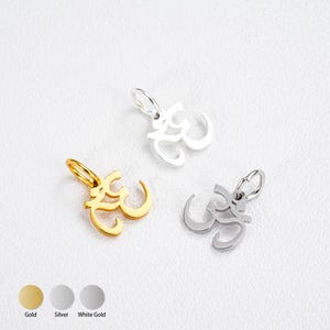 Op de afbeelding: Drie Om-symboolhangers in goud, zilver en witgoud. Elke hanger heeft een lusvormig ontwerp met een kleine lus voor een ketting. De hangers worden op een wit oppervlak weergegeven.