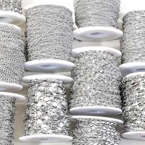 May include: An assortment of silver-colored chain spools, each wound around a white plastic bobbin. The chains vary in style, including delicate link, beaded, and sequin designs. The spools are arranged closely together, showcasing the variety of jewelry-making supplies.