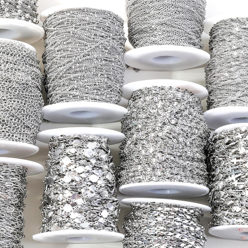 May include: An assortment of silver-colored chain spools, each wound around a white plastic bobbin. The chains vary in style, including delicate link, beaded, and sequin designs. The spools are arranged closely together, showcasing the variety of jewelry-making supplies.