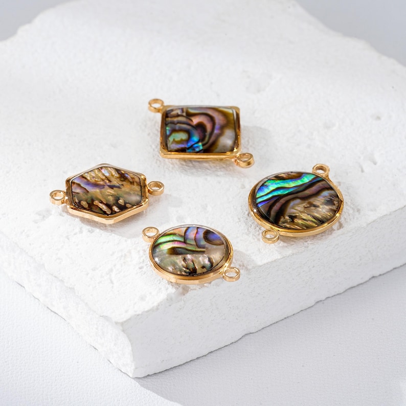 May include: Four gold-toned jewelry components with iridescent abalone shell inlays. The shapes include a rectangle, hexagon, and two circles. Each piece has small loops for attaching to other jewelry elements. The abalone shells display a range of colors.