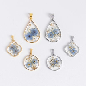 Puede incluir: Seis colgantes diferentes con flores prensadas en su interior. Los colgantes tienen forma de lágrima, círculo y trébol de cuatro hojas. Las flores son azules y blancas.