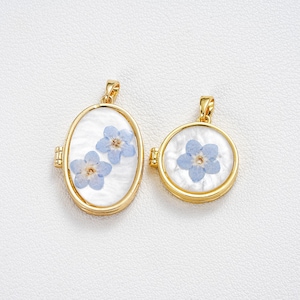 Può includere: Due ciondoli color oro con fiori blu non ti scordar di me. Uno è ovale, l'altro rotondo. Entrambi hanno uno sfondo bianco. I ciondoli sono probabilmente per collane o ciondoli.