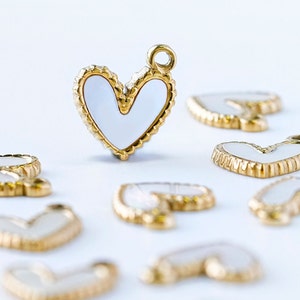 Puede incluir: Una colección de dijes de corazón de tono dorado con centros de esmalte blanco. Los dijes están esparcidos sobre una superficie blanca.