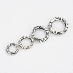 May include: Four silver-toned, textured round clasps of varying sizes arranged diagonally. Each clasp features a twisted rope design around the outer edge. The clasps are set against a white background.