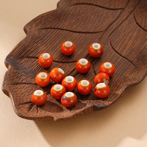 May include: A collection of orange ceramic beads with white centers, displayed on a carved wooden leaf-shaped tray. The beads have a glossy finish and are evenly spaced, showcasing their round shape and detailed craftsmanship. The tray's dark wood provides a contrasting backdrop.