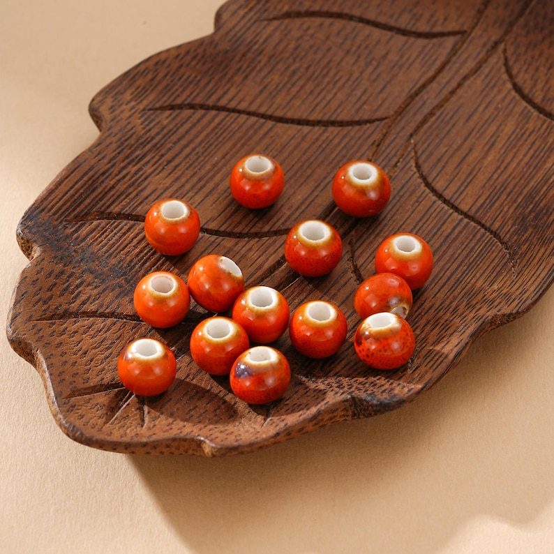 May include: A collection of orange ceramic beads with white centers, displayed on a carved wooden leaf-shaped tray. The beads have a glossy finish and are evenly spaced, showcasing their round shape and detailed craftsmanship. The tray's dark wood provides a contrasting backdrop.