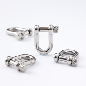 May include: Four shiny, silver D-shackles with screw pins against a white backdrop. The shackles are U-shaped with rounded ends and a threaded pin for secure closure. The screw pins have a textured, knurled design, typical of marine hardware.