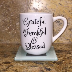 May include: White ceramic coffee mug with black text that reads "Grateful Thankful & Blessed". The mug is sitting on a clear glass coaster.