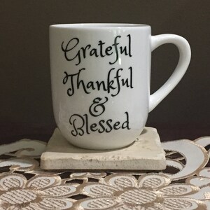 May include: White ceramic coffee mug with black lettering that reads "Grateful Thankful & Blessed". The mug is sitting on a beige coaster.