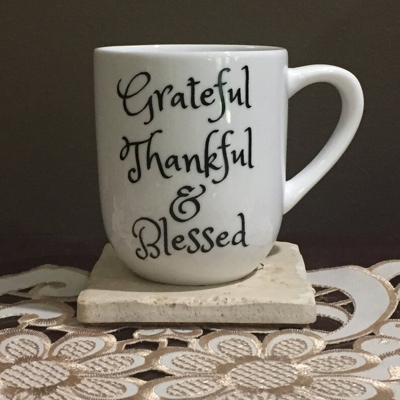 May include: White ceramic coffee mug with black lettering that reads "Grateful Thankful & Blessed". The mug is sitting on a beige coaster.