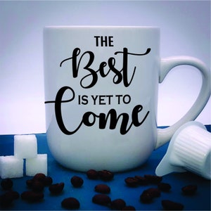 May include: White ceramic mug with the text "THE BEST IS YET TO COME" in black script. The mug is next to sugar cubes, coffee beans, and a creamer cup on a blue surface.
