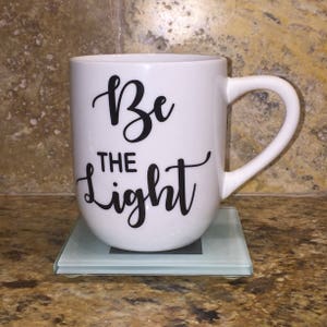 May include: White ceramic mug with a black inscription that reads "Be the light" in a cursive font. The mug is sitting on a light blue coaster.