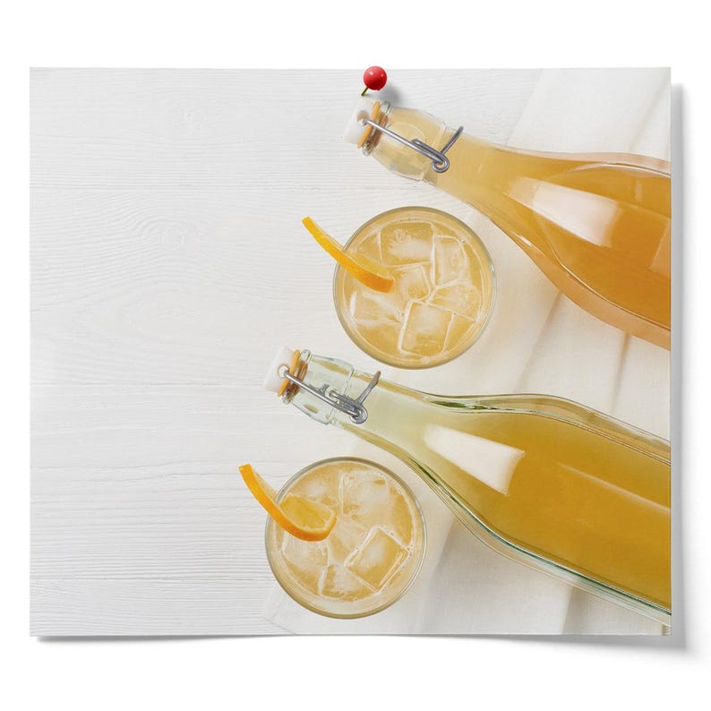 K&ouml;nnte beinhalten: Drei Glasflaschen gef&uuml;llt mit einer hellgelben Fl&uuml;ssigkeit, zwei der Flaschen liegen und eine steht aufrecht. Zwei klare Gl&auml;ser mit Eisw&uuml;rfeln und einer Orangenscheibe befinden sich neben den Flaschen.