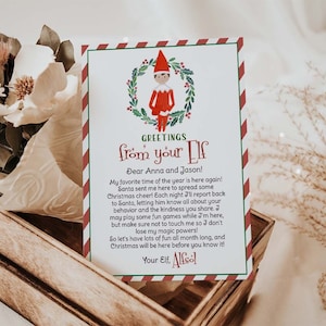 May include: A Christmas-themed card with a red and white striped border and the text "Greetings from your Elf." The card features an illustration of an elf and a wreath. The card is placed in a wooden crate with dried flowers.