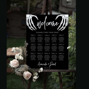 Puede incluir: Un cartel rectangular negro de distribuci&oacute;n de asientos para bodas con manos de esqueleto blancas en la parte superior, la palabra "welcome" en cursiva, el texto "PLEASE FIND YOUR SEAT", quince mesas con nombres y "Amanda + David" con "10.31.XX".