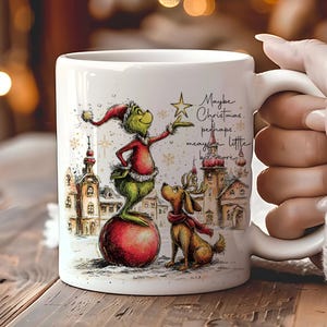 May include: White ceramic mug with a festive Christmas design. The illustration shows a green character in a Santa hat, a dog with a scarf, and the text "Maybe Christmas..." The mug is held by a person.