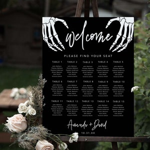 Puede incluir: Un cartel negro de distribuci&oacute;n de asientos para bodas con manos de esqueleto blancas formando un coraz&oacute;n en la parte superior, el texto "Welcome", "Please find your seat", "Amanda + David" y la fecha "10.31.XX" con una lista de mesas y nombres.