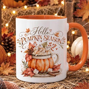 May include: A white ceramic mug with an orange interior and handle, featuring a graphic of a pumpkin-shaped cup filled with whipped cream and marshmallows, surrounded by autumn leaves and the text Hello PUMPKIN SEASON in brown script.