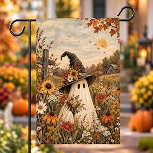 May include: A rectangular garden flag featuring a white ghost wearing a tall, dark, pointed hat adorned with sunflowers. The ghost stands in a field of autumn wildflowers, including orange and yellow blooms, with a wooden cross and a small house in the distance under a pale sky with a full moon and flying birds.