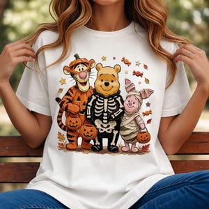 May include: A white t-shirt featuring a graphic of Tigger dressed as a pumpkin, Winnie the Pooh wearing a skeleton costume, and Piglet dressed as a mummy. The characters are surrounded by small orange candies and autumn leaves.