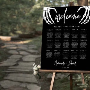 Puede incluir: Un cartel rectangular negro para la distribuci&oacute;n de asientos de boda con manos de esqueleto blancas que forman un coraz&oacute;n en la parte superior. El texto dice welcome, please find your seat, table 1 al 15 con nombres y Amanda + David con la fecha 10.31.XX.