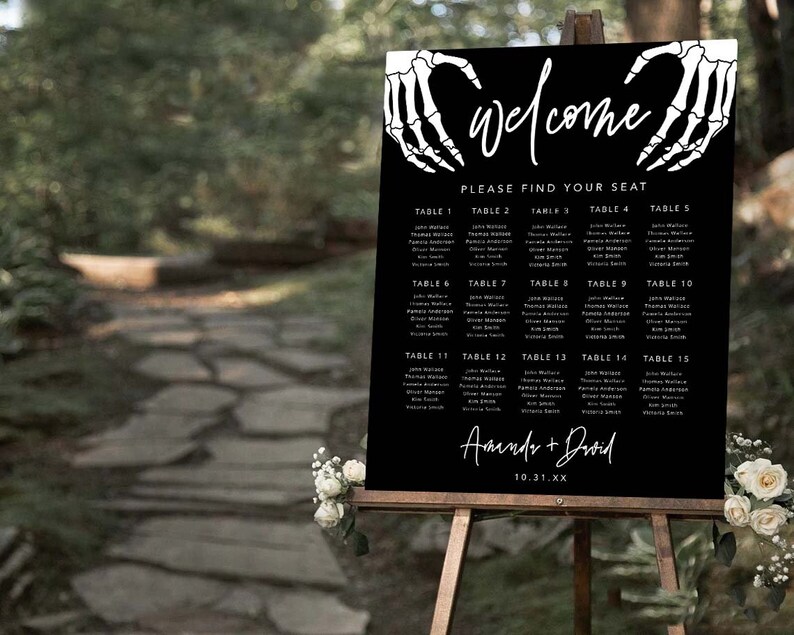 Puede incluir: Un cartel rectangular negro para la distribuci&oacute;n de asientos de boda con manos de esqueleto blancas que forman un coraz&oacute;n en la parte superior. El texto dice welcome, please find your seat, table 1 al 15 con nombres y Amanda + David con la fecha 10.31.XX.