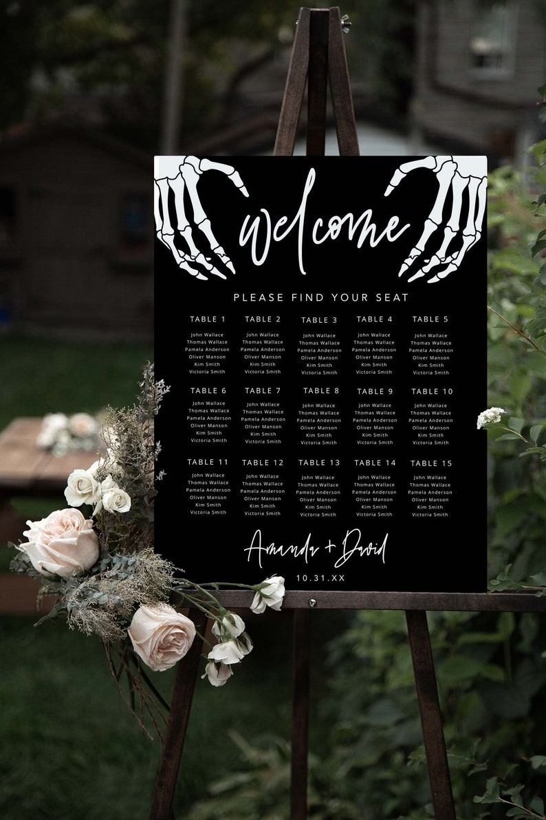 Puede incluir: Un cartel negro de distribuci&oacute;n de asientos para bodas con manos de esqueleto blancas formando un coraz&oacute;n en la parte superior, el texto "Welcome", "Please find your seat", "Amanda + David" y la fecha "10.31.XX" con una lista de mesas y nombres.