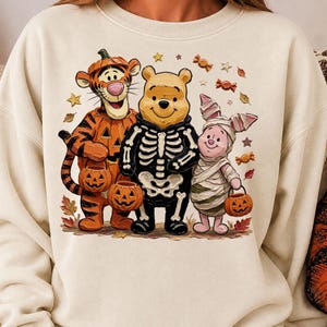 May include: A cream-colored crewneck sweatshirt featuring a graphic print of Tigger dressed as a pumpkin, Winnie the Pooh in a skeleton costume, and Piglet wrapped as a mummy. They are holding jack-o'-lantern buckets surrounded by autumn leaves.