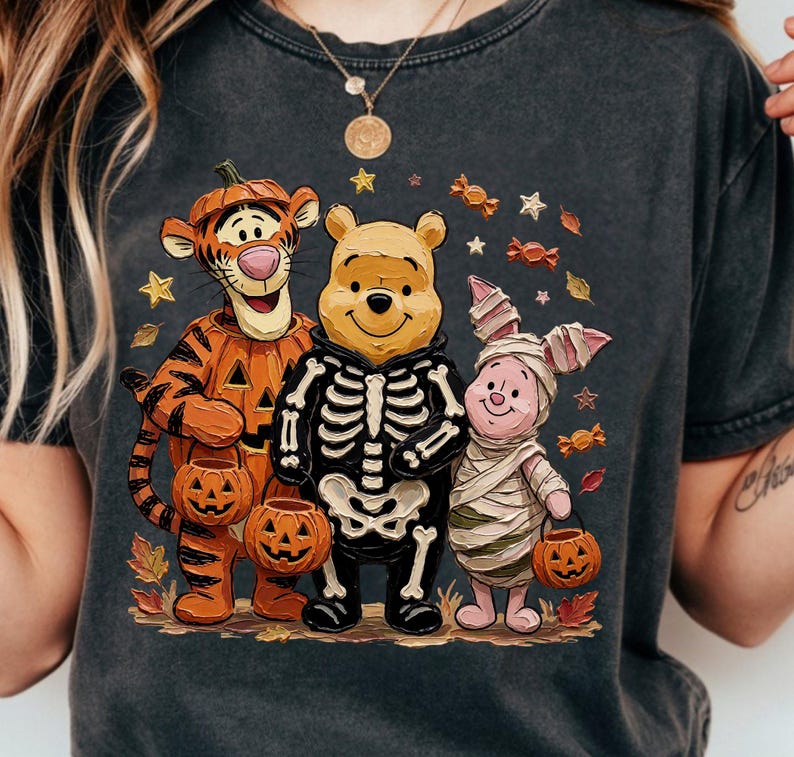 May include: A charcoal gray t-shirt featuring a graphic of Tigger dressed as a pumpkin, Winnie the Pooh wearing a skeleton costume, and Piglet wrapped as a mummy. The characters are holding small jack-o'-lantern buckets amidst scattered candy and autumn leaves.