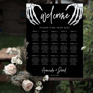 Puede incluir: Un cartel negro de distribuci&oacute;n de asientos para bodas con manos de esqueleto blancas que forman un coraz&oacute;n en la parte superior. El texto dice "welcome", "PLEASE FIND YOUR SEAT", "Amanda + David" y "10.31.XX", con mesas numeradas del 1 al 15.