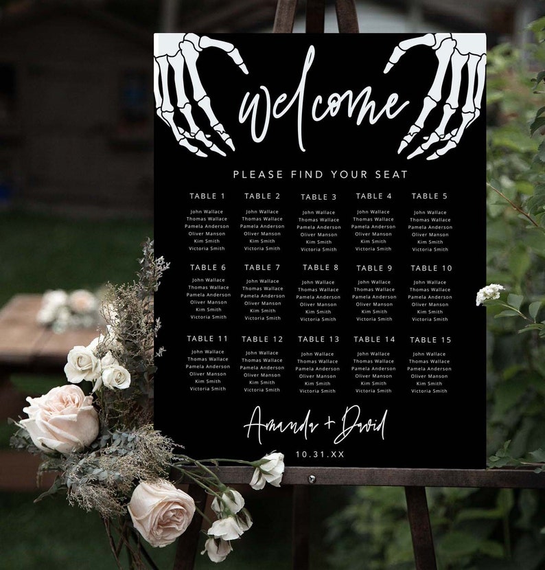 Puede incluir: Un cartel negro de distribuci&oacute;n de asientos para bodas con manos de esqueleto blancas que forman un coraz&oacute;n en la parte superior. El texto dice "welcome", "PLEASE FIND YOUR SEAT", "Amanda + David" y "10.31.XX", con mesas numeradas del 1 al 15.