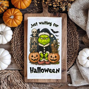 May include: A white waffle-weave kitchen towel with a Halloween design. The towel features a green character in a black coat, two jack-o'-lanterns, a skeleton, and the text "Just waiting for Halloween".