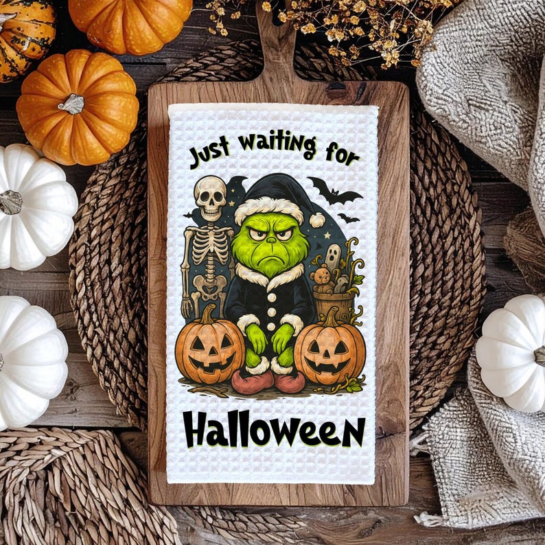 May include: A white waffle-weave kitchen towel with a Halloween design. The towel features a green character in a black coat, two jack-o'-lanterns, a skeleton, and the text "Just waiting for Halloween".