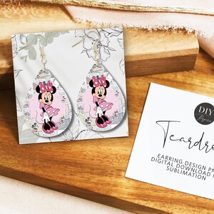 Op de afbeelding: Druppelvormige oorbellen met een Minnie Mouse-ontwerp. De oorbellen hebben een roze en zwart kleurenschema, met een cartoonafbeelding van Minnie Mouse. De oorbellen worden gepresenteerd op een witte kaart met bloemaccenten.