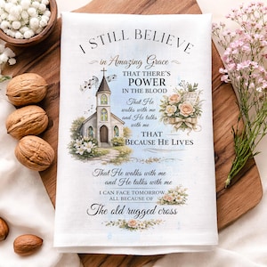 May include: White linen kitchen towel with the text "I Still Believe" and a church illustration. The towel also features floral accents and inspirational text. The towel is on a wooden cutting board.