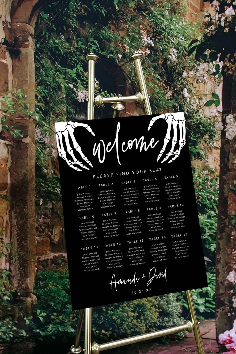 Puede incluir: Un cartel rectangular negro para la distribuci&oacute;n de asientos de boda con texto blanco y gr&aacute;ficos de manos de esqueleto. El cartel dice Welcome, Please Find Your Seat, enumera las mesas 1 a 15 con nombres y los nombres Amanda + David con 10.31.XX.