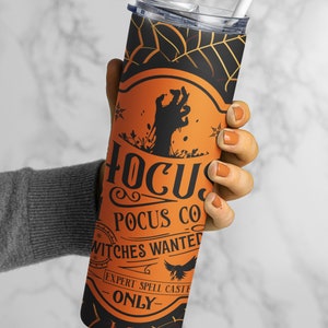 May include: A tall, black and orange Halloween-themed tumbler with a lid and straw. The tumbler features the text "Hocus Pocus Co" and "Witches Wanted" with a zombie hand graphic and spiderweb design. A spooky accessory for Halloween.