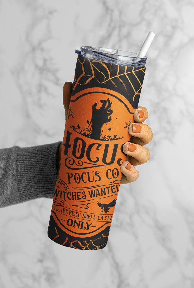 May include: A tall, black and orange Halloween-themed tumbler with a lid and straw. The tumbler features the text "Hocus Pocus Co" and "Witches Wanted" with a zombie hand graphic and spiderweb design. A spooky accessory for Halloween.