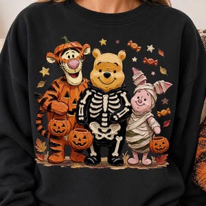 May include: A black crewneck sweatshirt featuring a graphic of Tigger dressed as a pumpkin, Winnie the Pooh wearing a skeleton costume, and Piglet wrapped as a mummy. The characters are holding small jack-o'-lantern buckets amidst falling autumn leaves, stars, and wrapped candies.