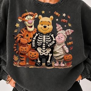 May include: A charcoal gray crewneck sweatshirt featuring a graphic print of Tigger dressed as a pumpkin, Winnie the Pooh in a skeleton costume, and Piglet wrapped as a mummy, all holding jack-o'-lantern buckets amidst scattered autumn leaves.
