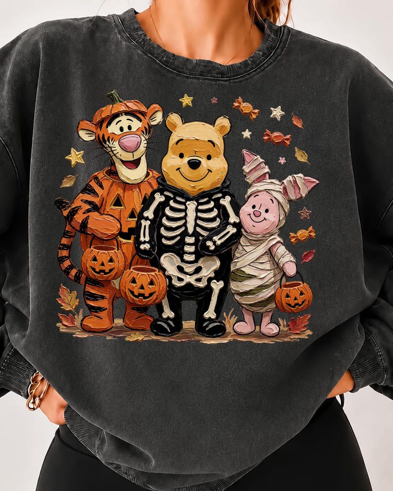 May include: A charcoal gray crewneck sweatshirt featuring a graphic print of Tigger dressed as a pumpkin, Winnie the Pooh in a skeleton costume, and Piglet wrapped as a mummy, all holding jack-o'-lantern buckets amidst scattered autumn leaves.