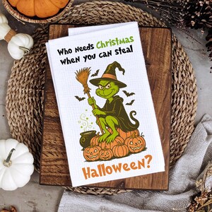 May include: White kitchen towel featuring a Grinch-themed Halloween design. The Grinch, in a witch hat, holds a broom, with bats and pumpkins around him. Text reads "Who needs Christmas when you can steal Halloween?"